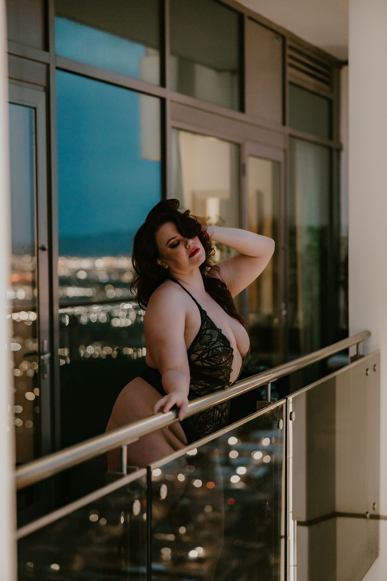 Woman in lace lingerie leans against balcony railing during sunset with cityscape view.