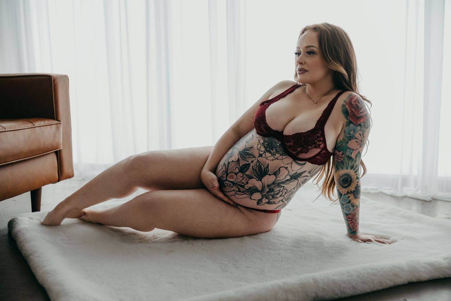 A person in black lingerie poses on a white couch in a softly lit room with sheer curtains.