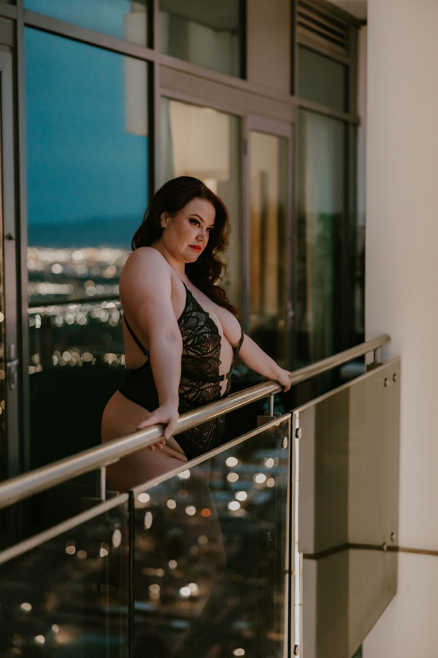 Woman in black dress leans against balcony railing at night with city lights glowing in background.