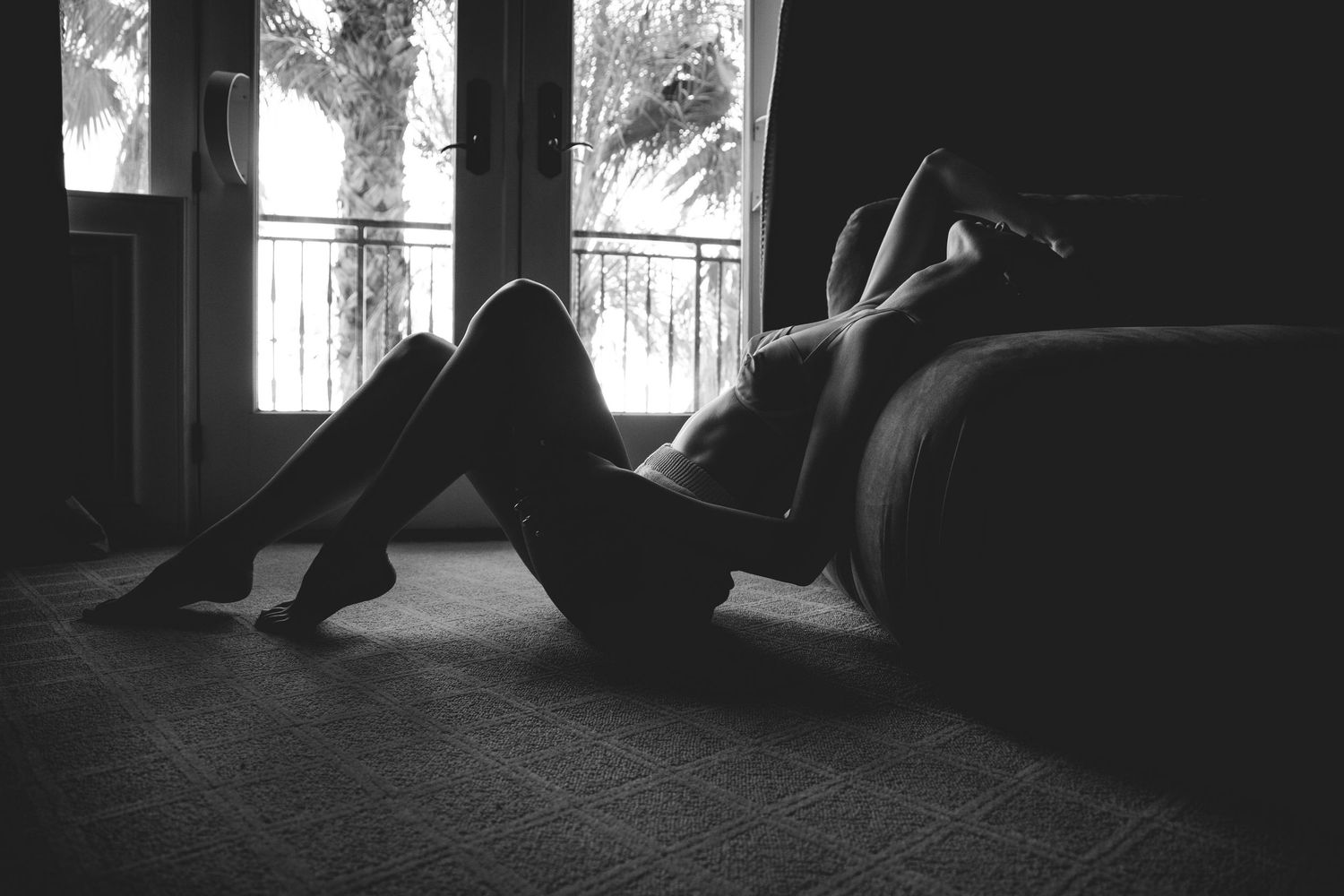 A black and white photo shows a person lying on a dark carpeted floor near a window in a dimly lit room.