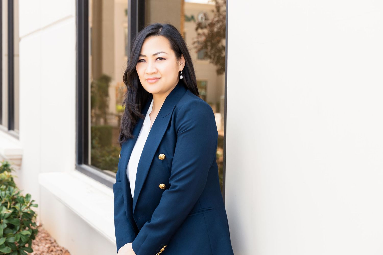 Professional woman in navy blazer smiling outdoors against white wall in corporate business portrait photo.