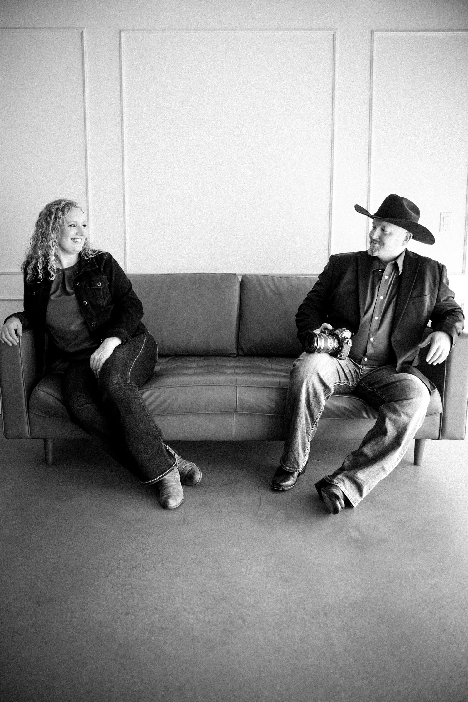 Black and white photo of a man in cowboy hat and woman sitting on opposite ends of a gray sofa, smiling at each other.