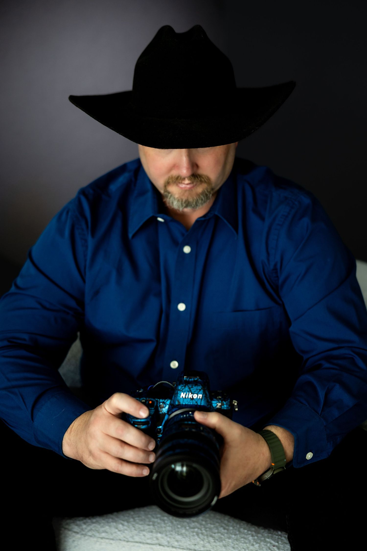 Man in black cowboy hat and blue shirt holding a Nikon camera, portrait photography with dramatic dark background.
