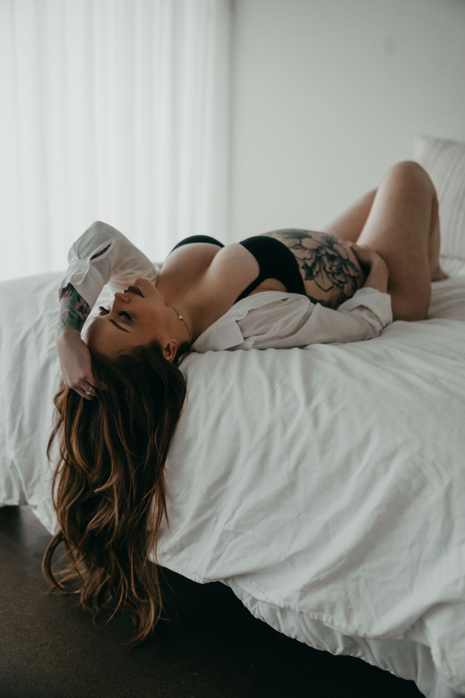 A woman in lingerie poses sensually on white bedding in a moody boudoir photoshoot series.