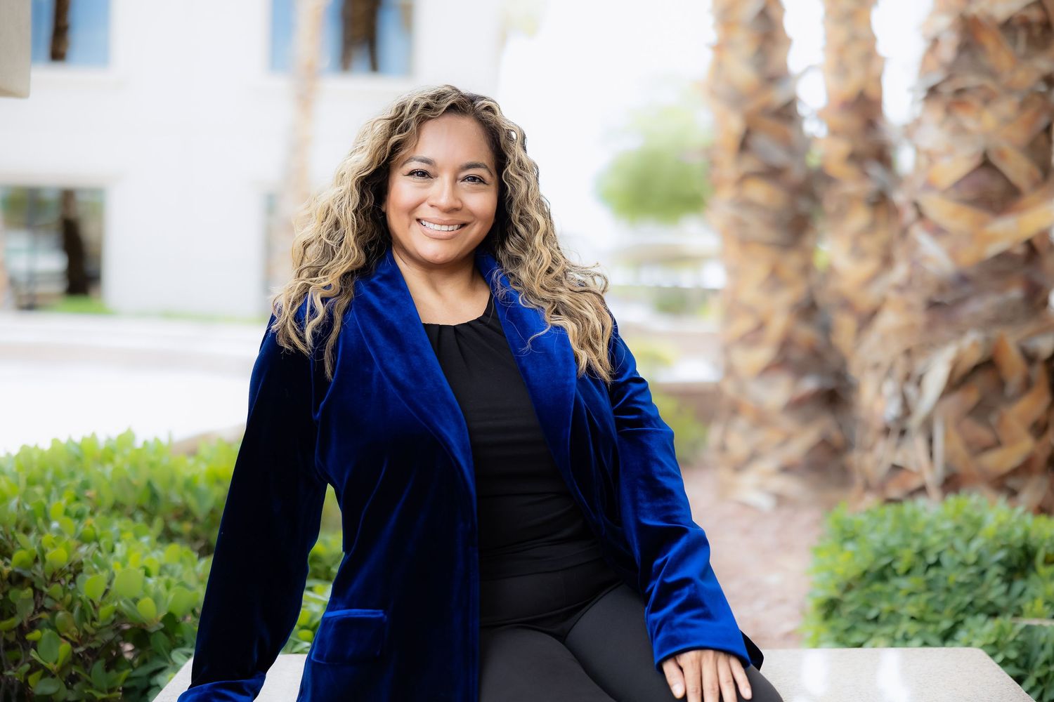 Woman in blue velvet blazer smiling outdoors near palm trees and greenery in a professional portrait.