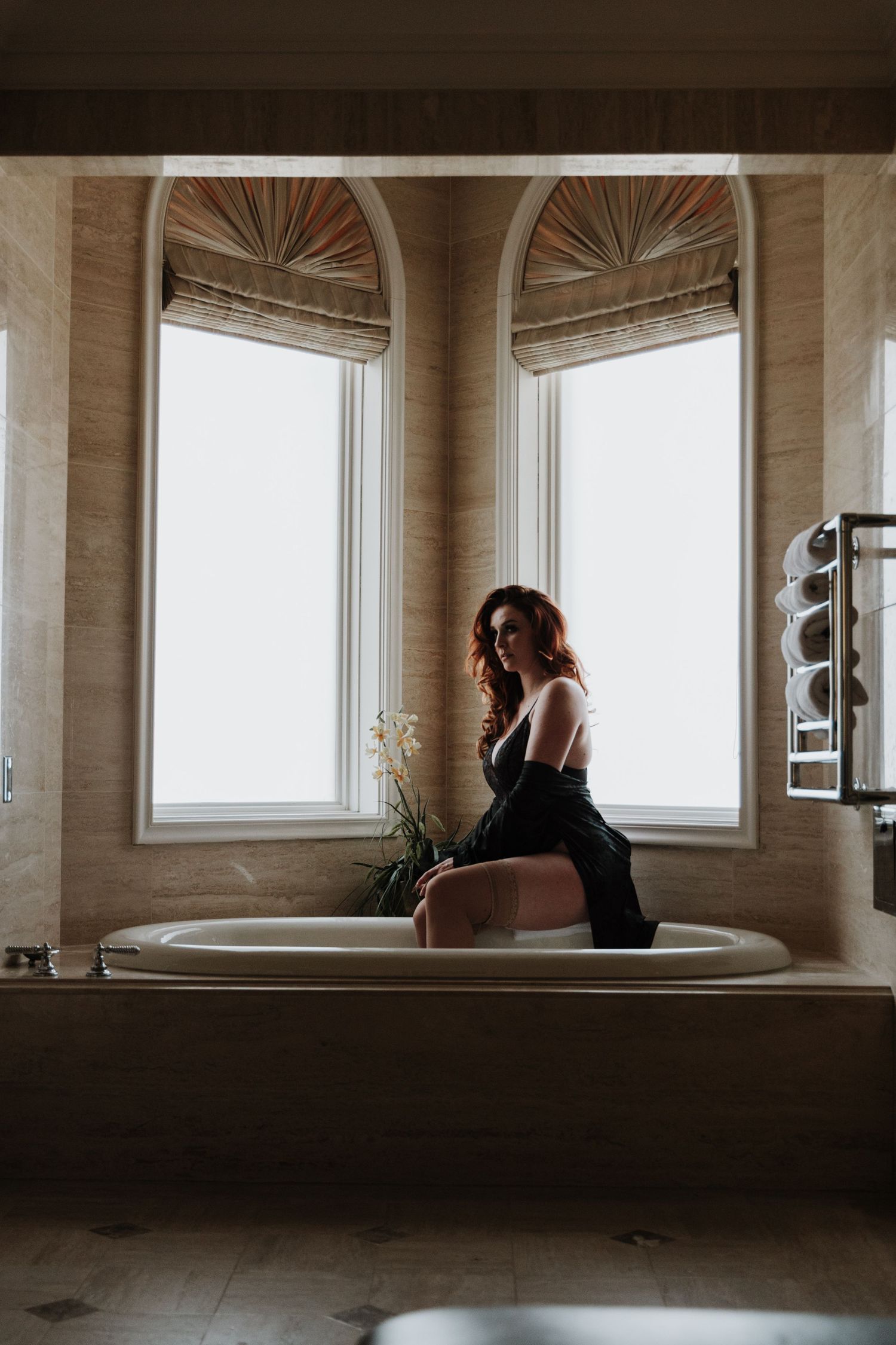 Moody boudoir photo of figure sitting on bathtub edge near arched windows.