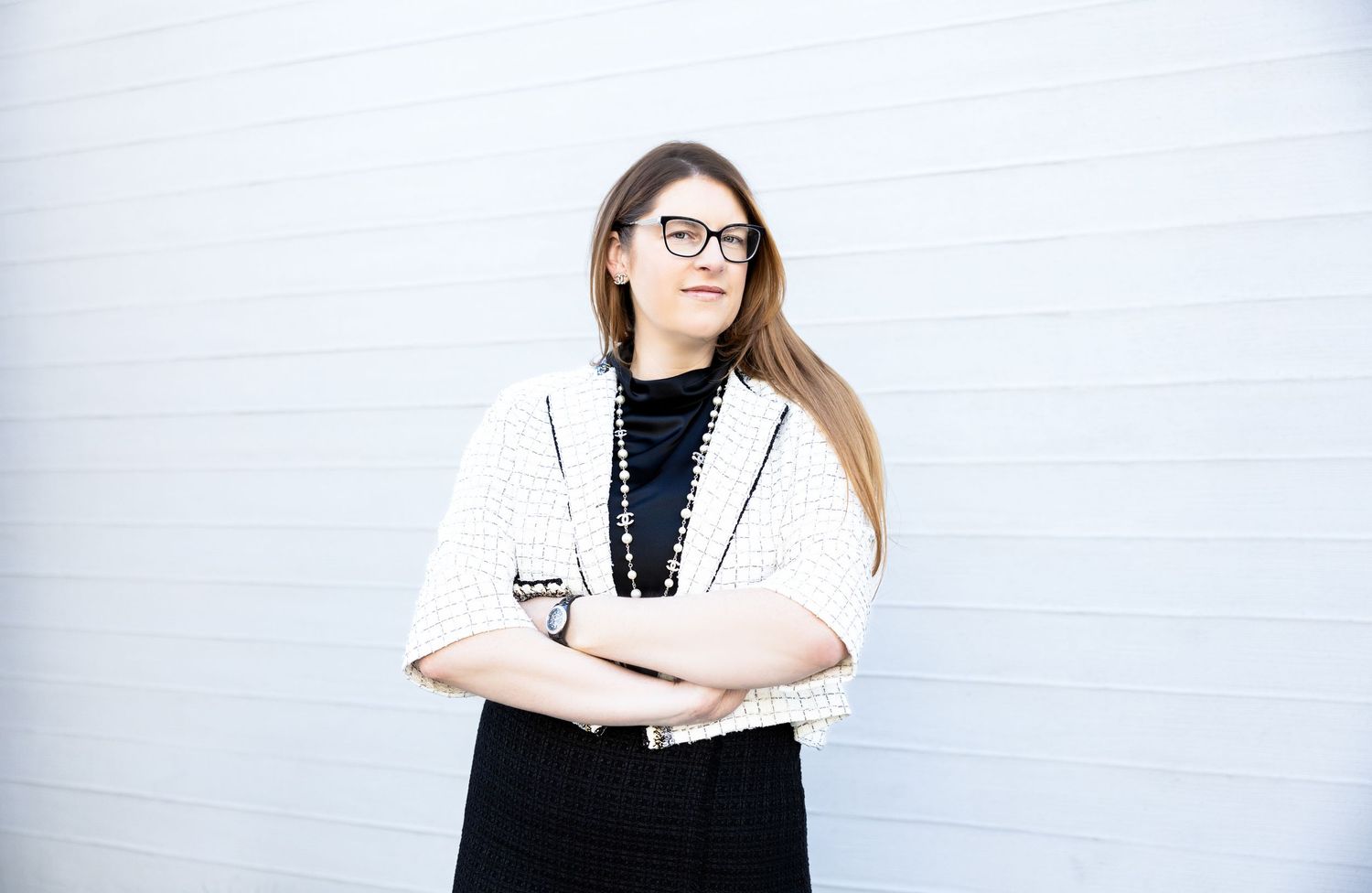 Woman with glasses and long hair poses confidently with arms crossed, wearing a white top and black necklace.