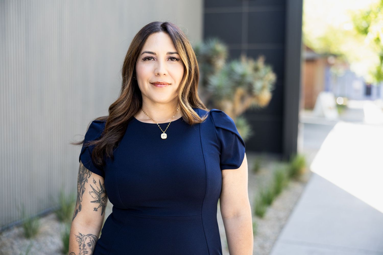 A professional woman in a navy blue dress with a tattoo on her arm poses outdoors in a modern setting.