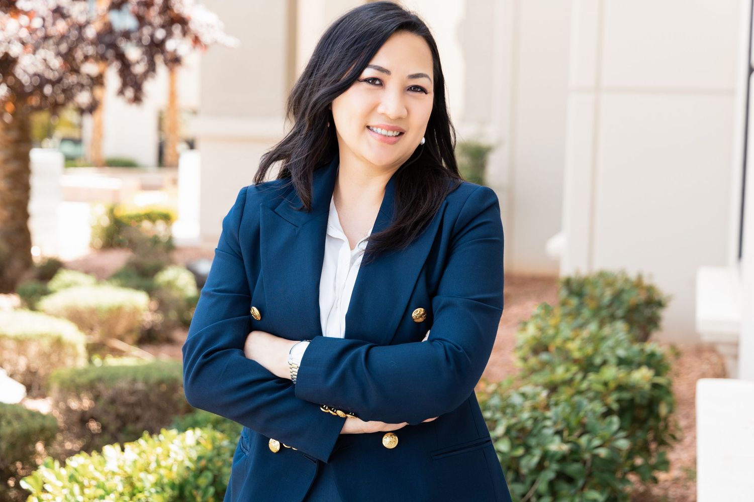 Professional woman in navy blazer smiling with arms crossed outdoors in a corporate setting.