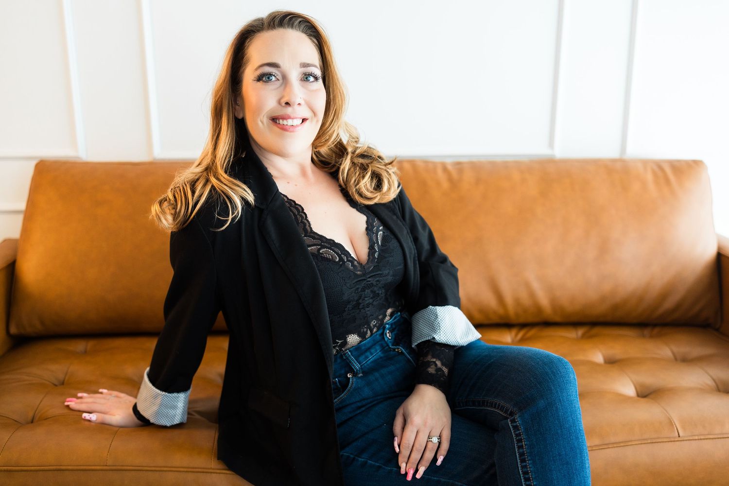 Woman in black blazer and jeans sitting relaxed on a brown leather sofa, smiling confidently.