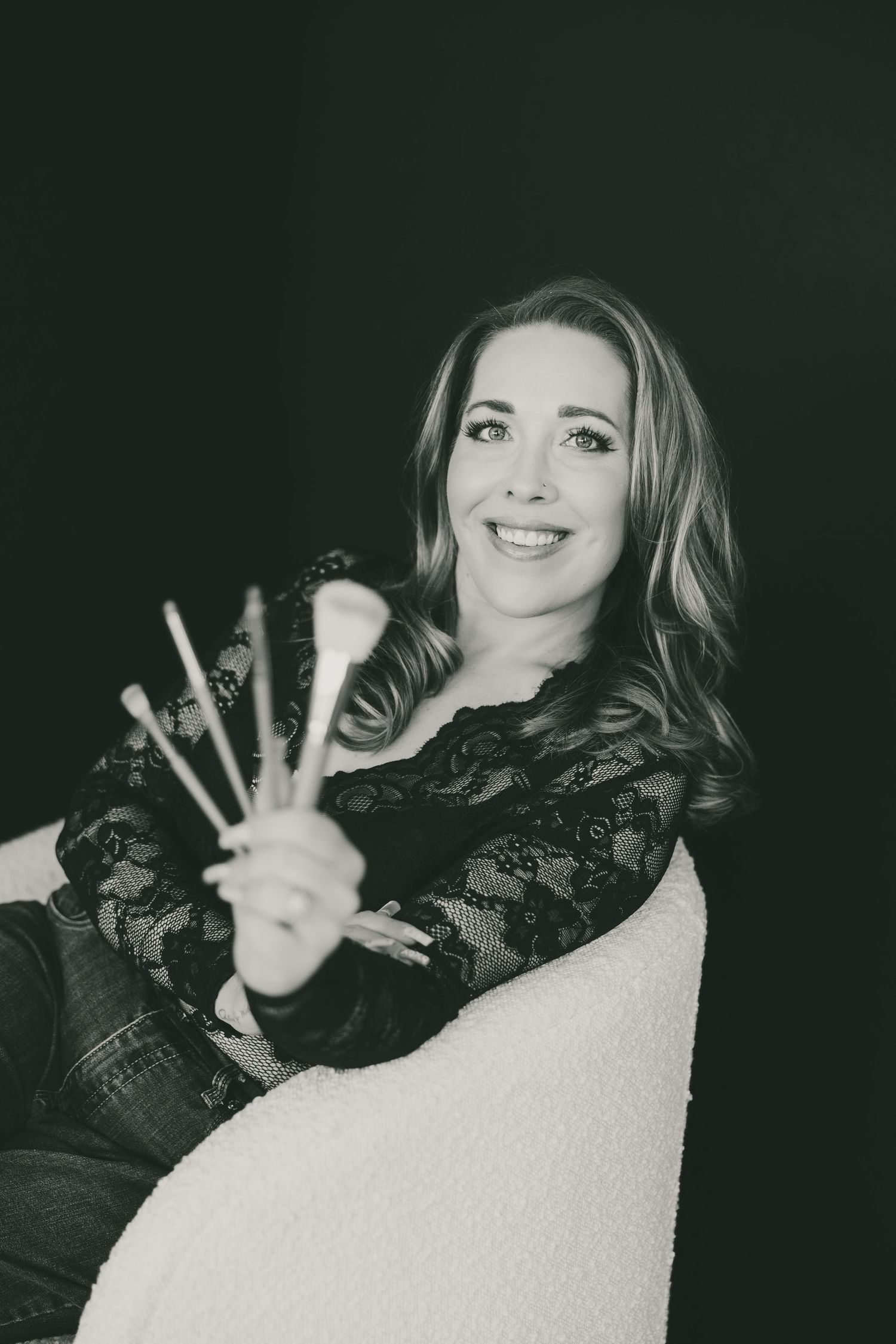 Black and white photo of smiling woman holding makeup brushes while seated in a white chair against dark background.