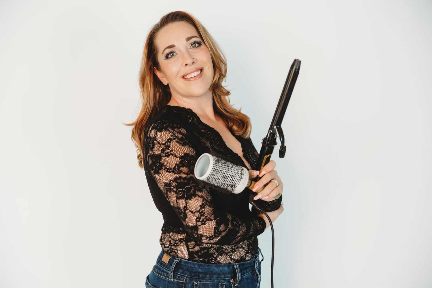 Woman in black lace top holding curling iron and brush, smiling against white background.
