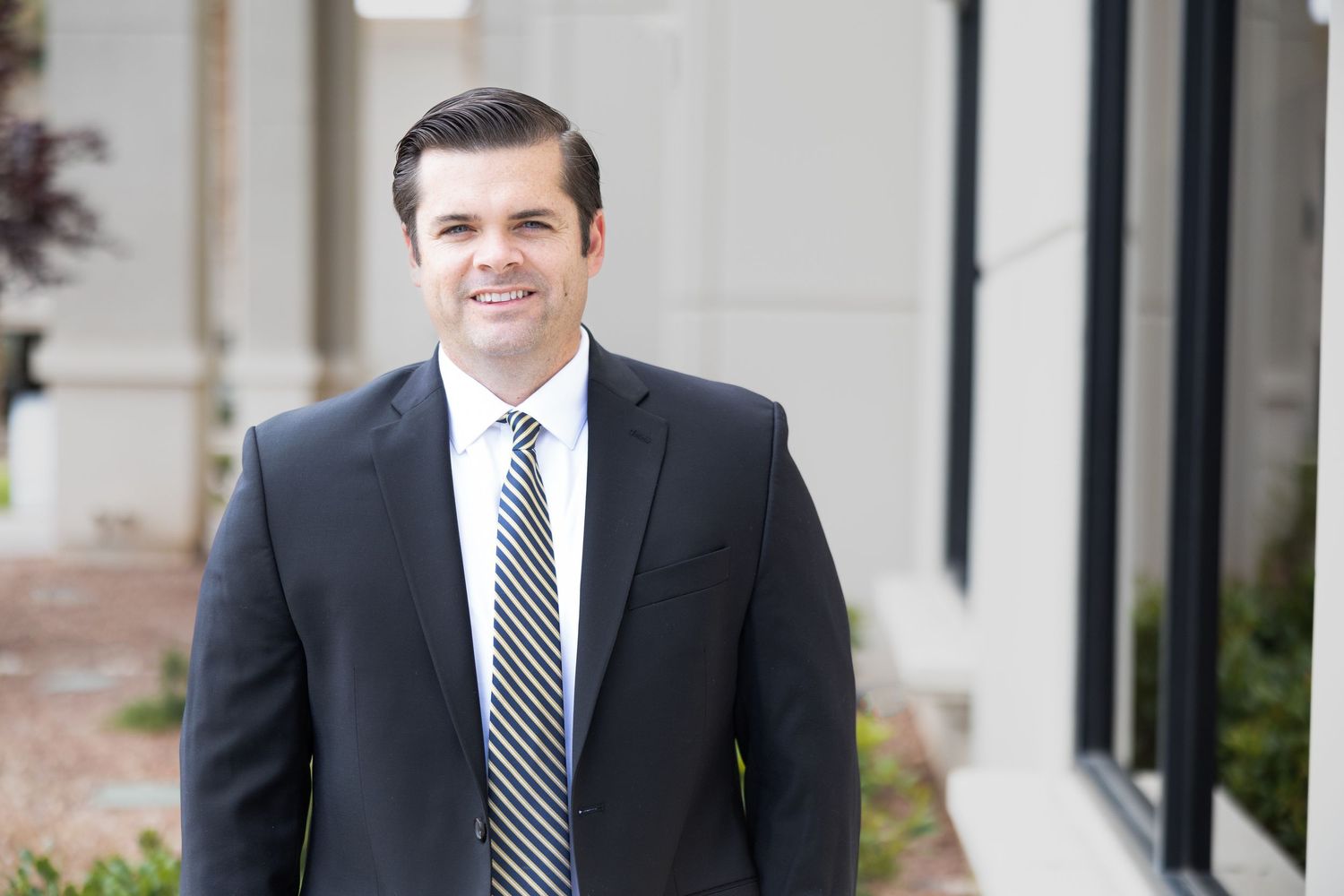 Professional businessman in dark suit and striped tie smiling outdoors near modern building columns.