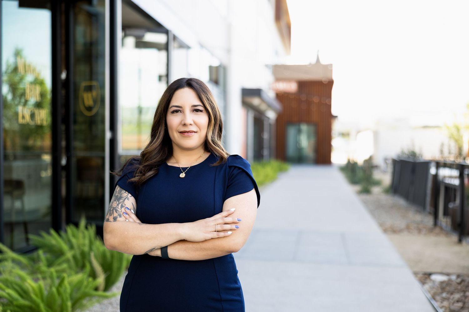Professional woman in navy dress with tattoo stands arms crossed outside modern building.