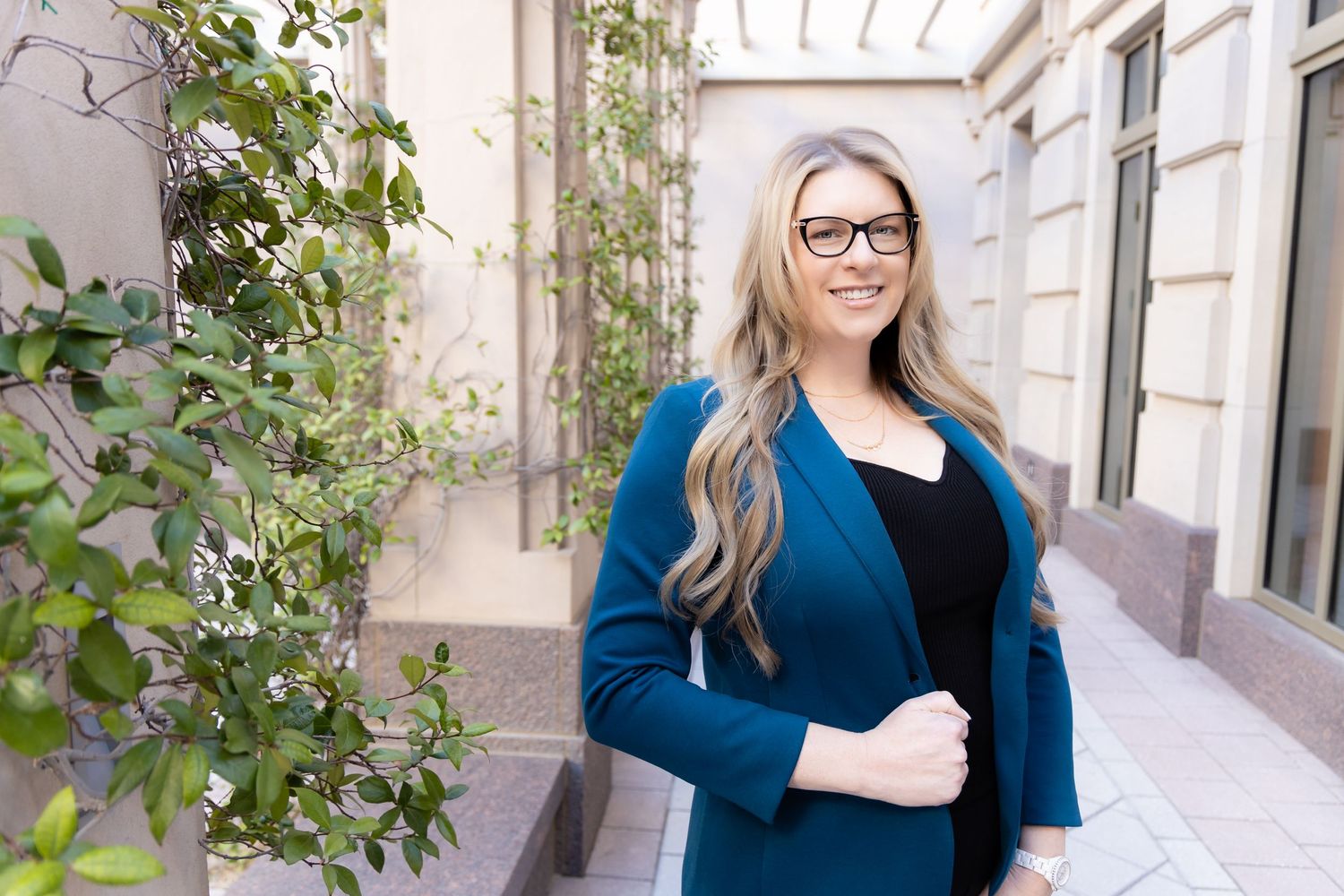 Professional woman in teal blazer and glasses smiling in elegant outdoor corridor with lush greenery.