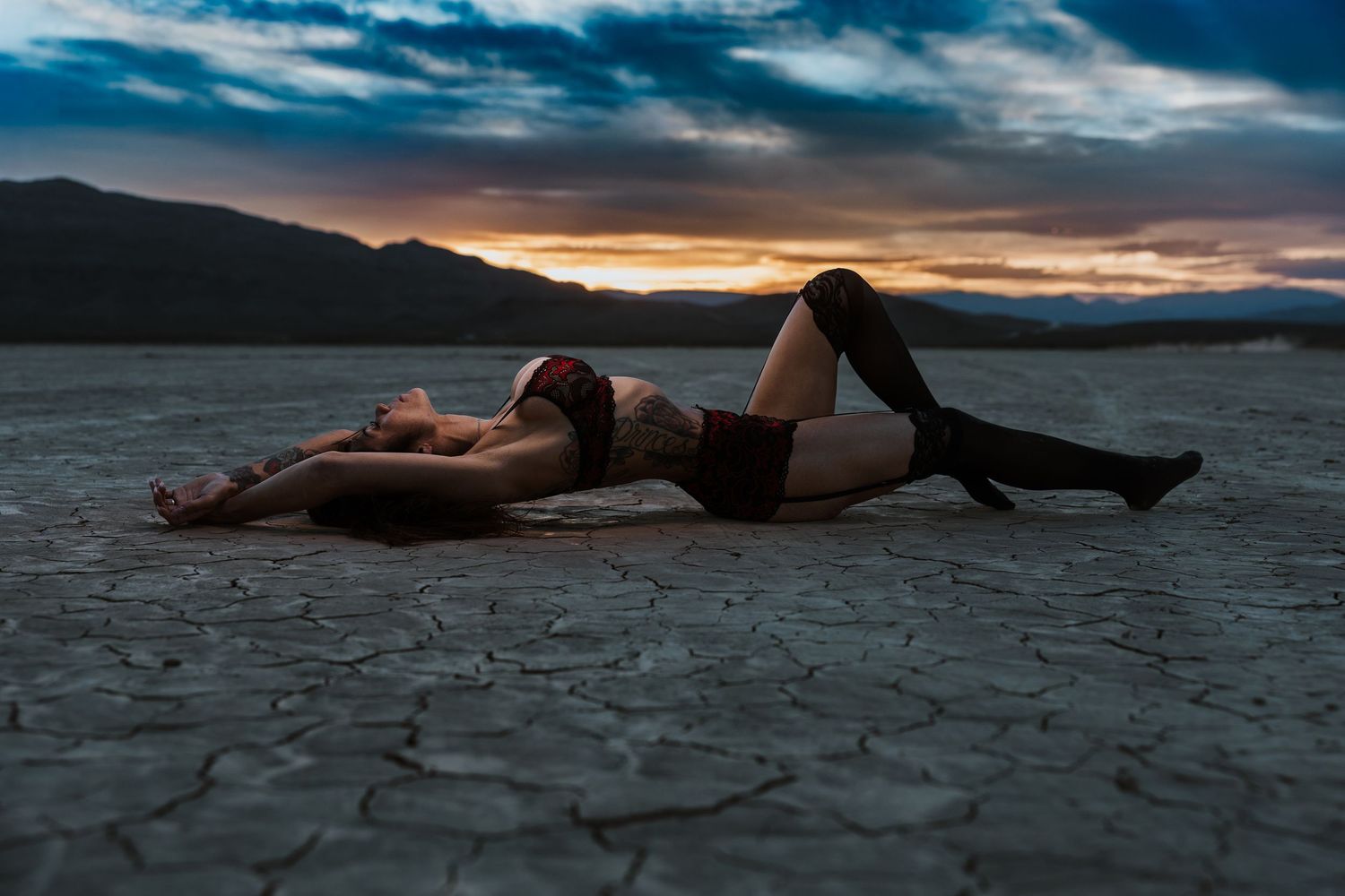 A person lies on cracked desert ground against a dramatic sunset sky with clouds and mountains in the distance.