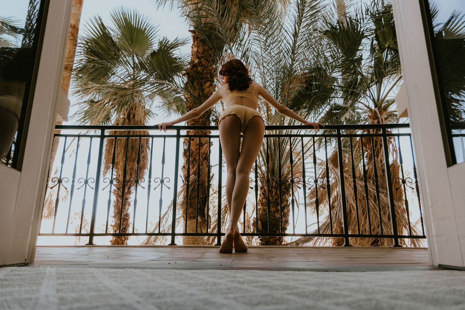 Person in beige outfit stretches on a balcony with palm trees and iron railing during golden hour.