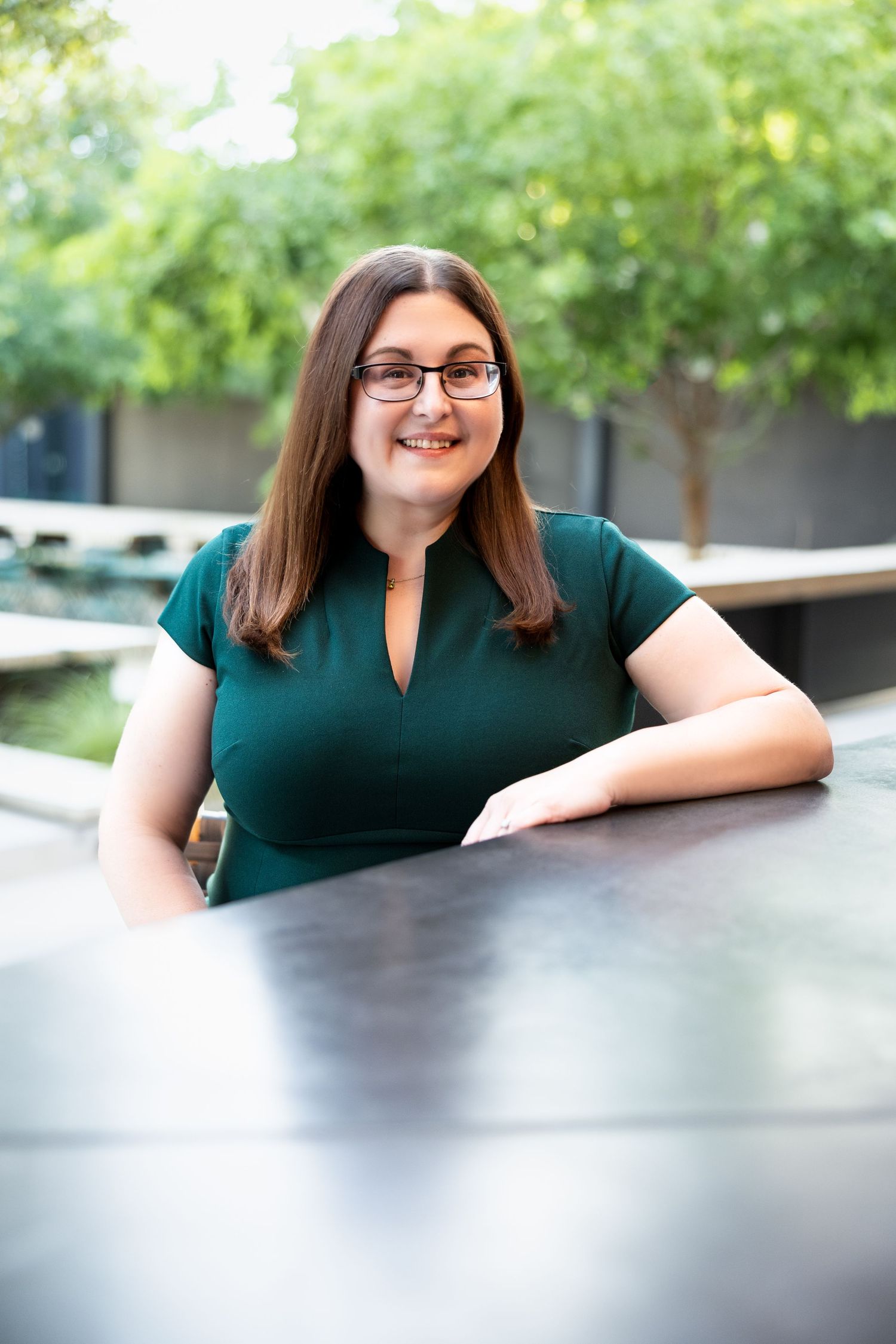 Woman with glasses in green top smiling outdoors, seated at a dark table with green trees in background.