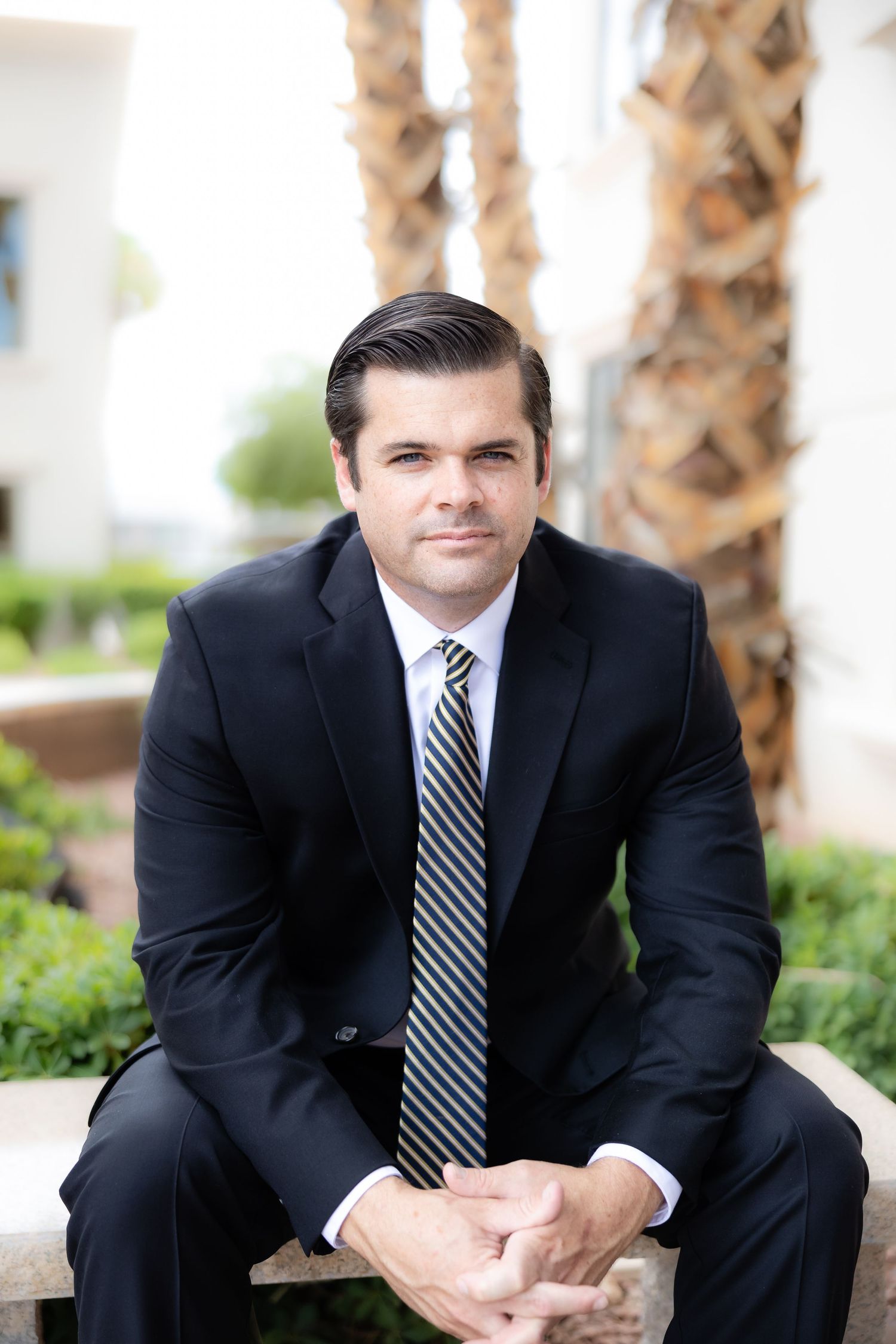 Professional man in black suit and striped tie sits outdoors with palm trees, ideal for business or legal profile photo.