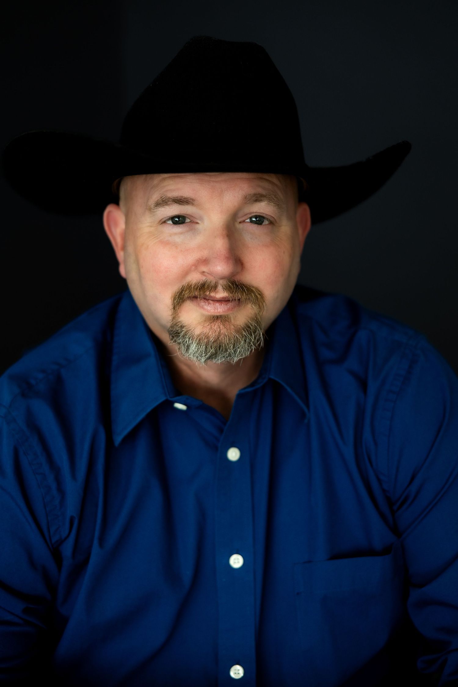 A middle-aged man with a goatee wearing a black cowboy hat and blue button-up shirt poses for a portrait.