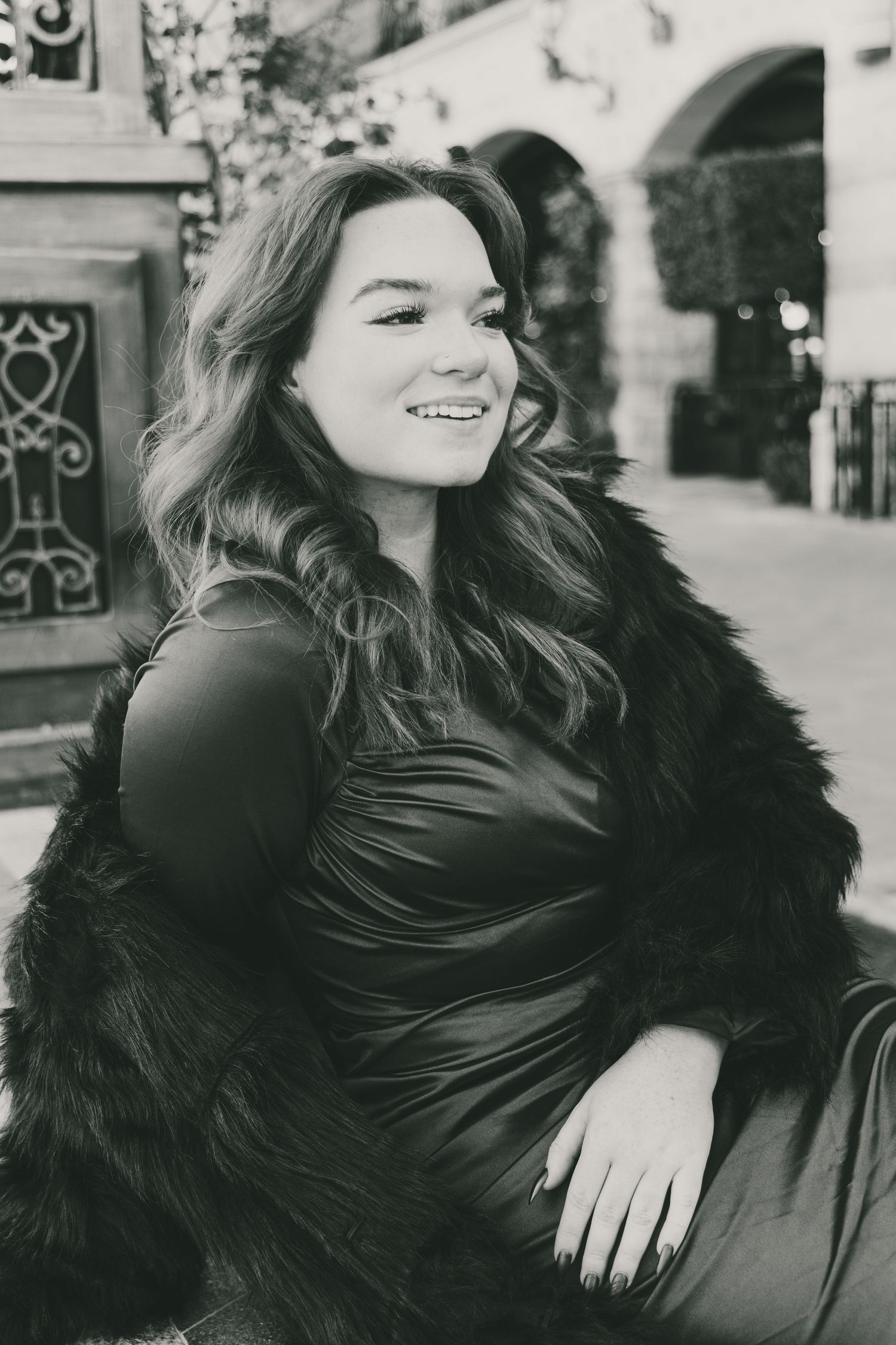 Black and white photo of a smiling young woman in a fur coat and satin dress posing outdoors.