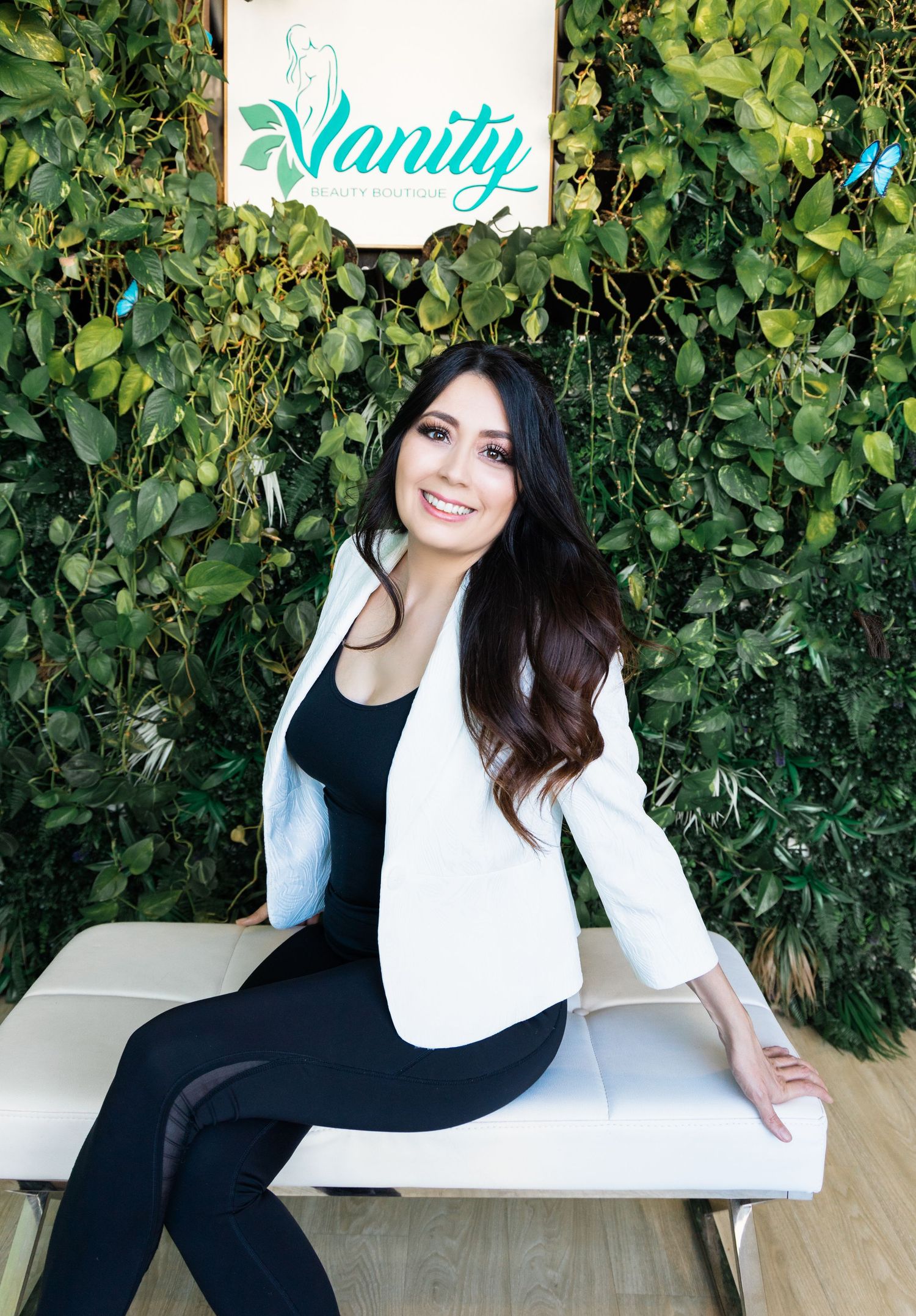 Woman in white blazer and black outfit smiling at Vanity salon, posing in front of a lush green plant wall.