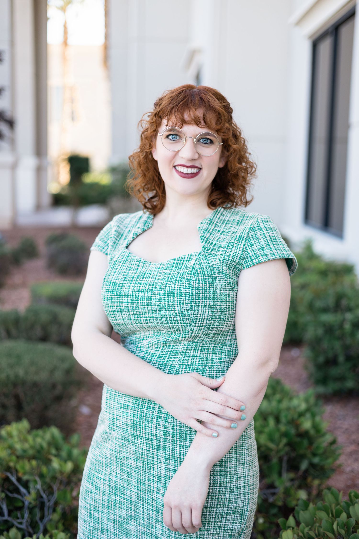 Smiling redhead woman in green tweed dress poses outdoors near shrubs and building columns.