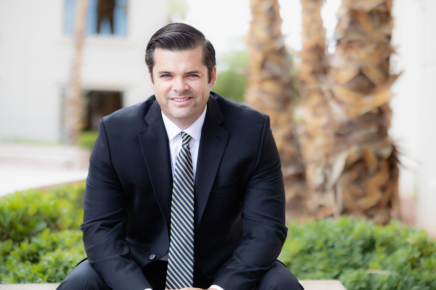 Professional businessman in dark suit and striped tie smiling outdoors near palm trees and green hedges.