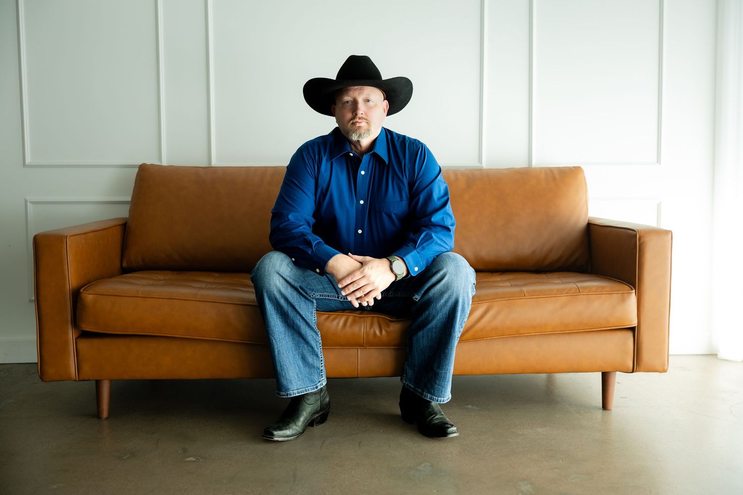 Man in black cowboy hat and blue shirt sitting on brown leather sofa against white wall.