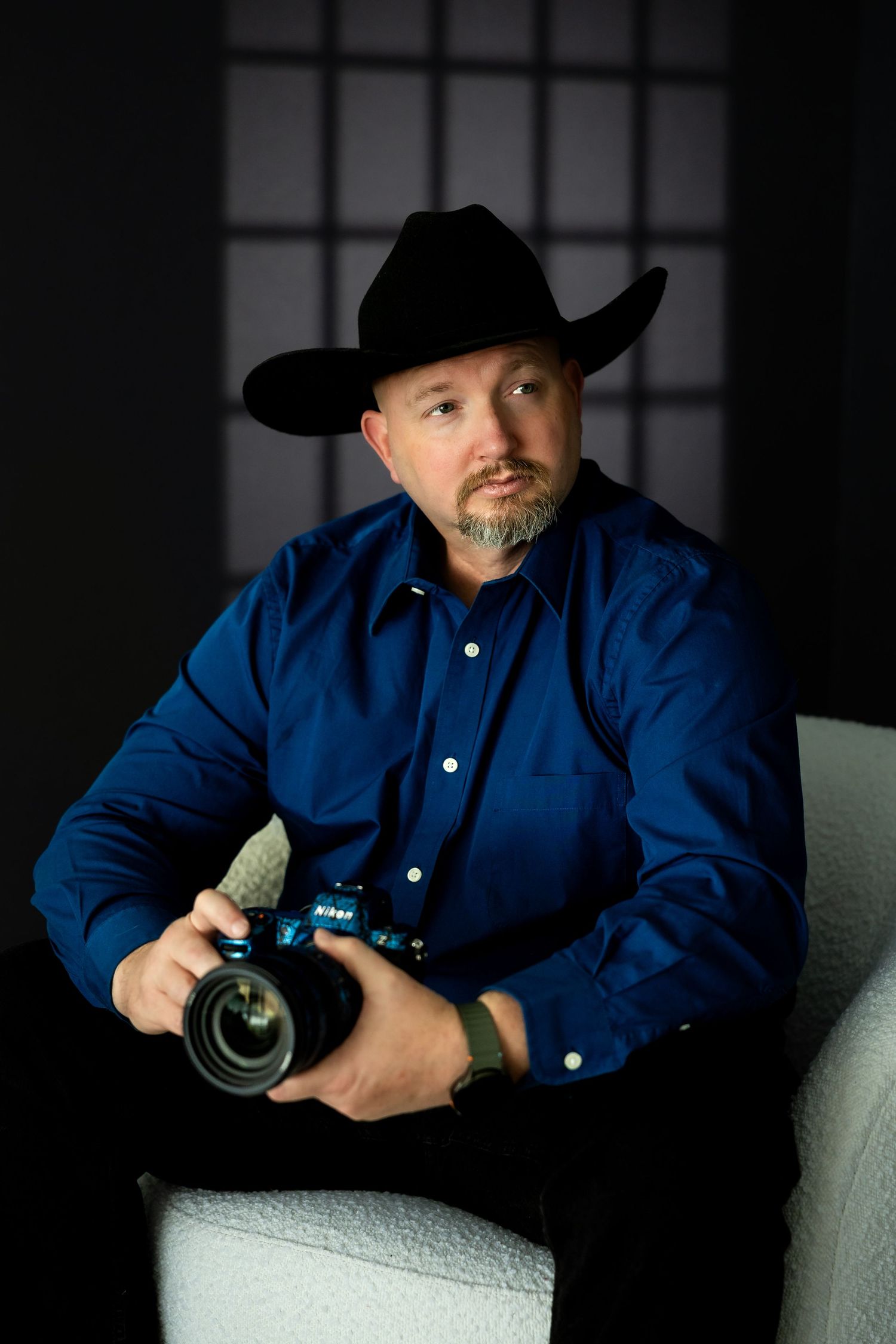 Man in black cowboy hat and blue shirt holding a Nikon camera, seated in a white chair against a dark background.