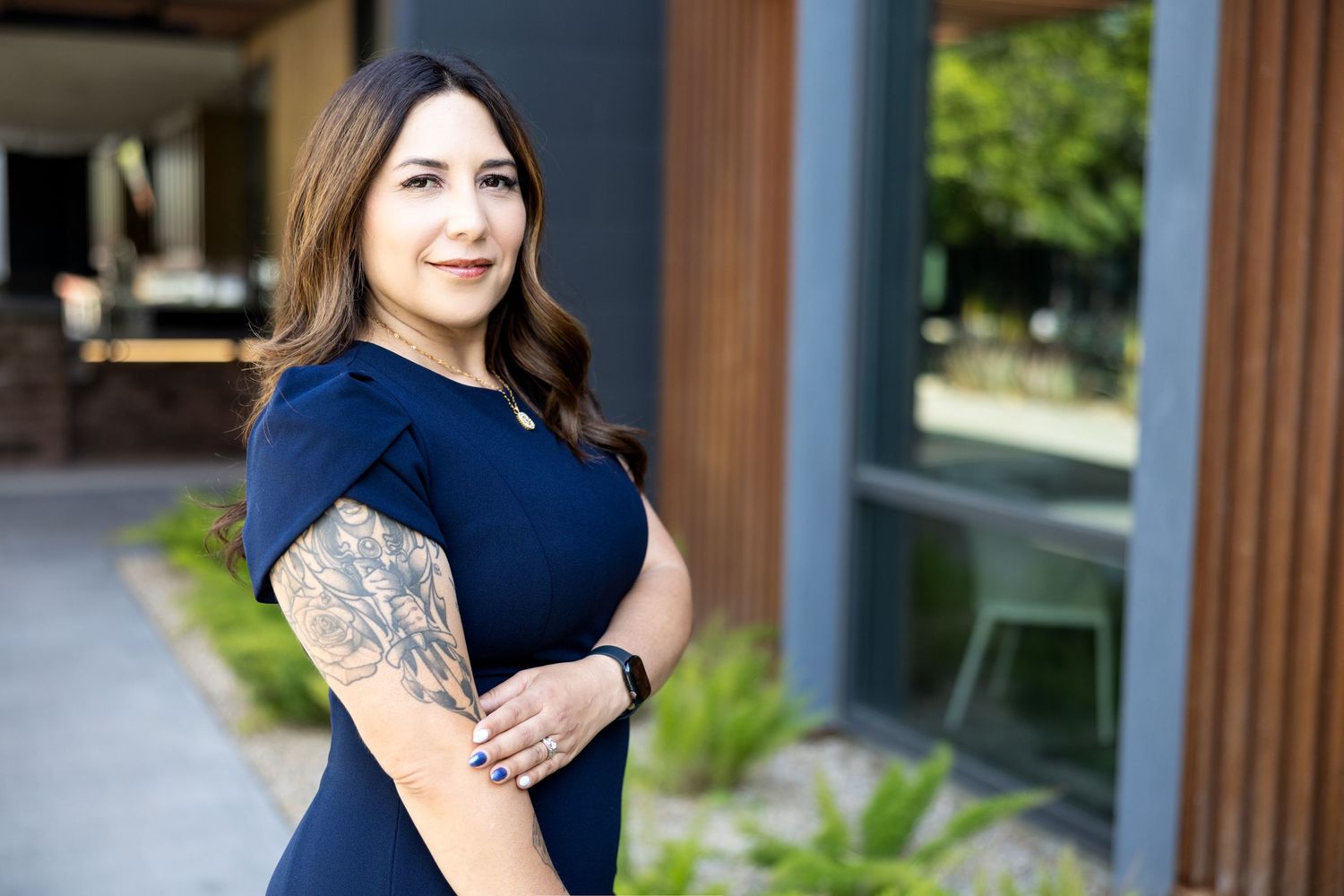 Professional woman in navy dress with floral arm tattoo posing confidently outdoors at modern building.
