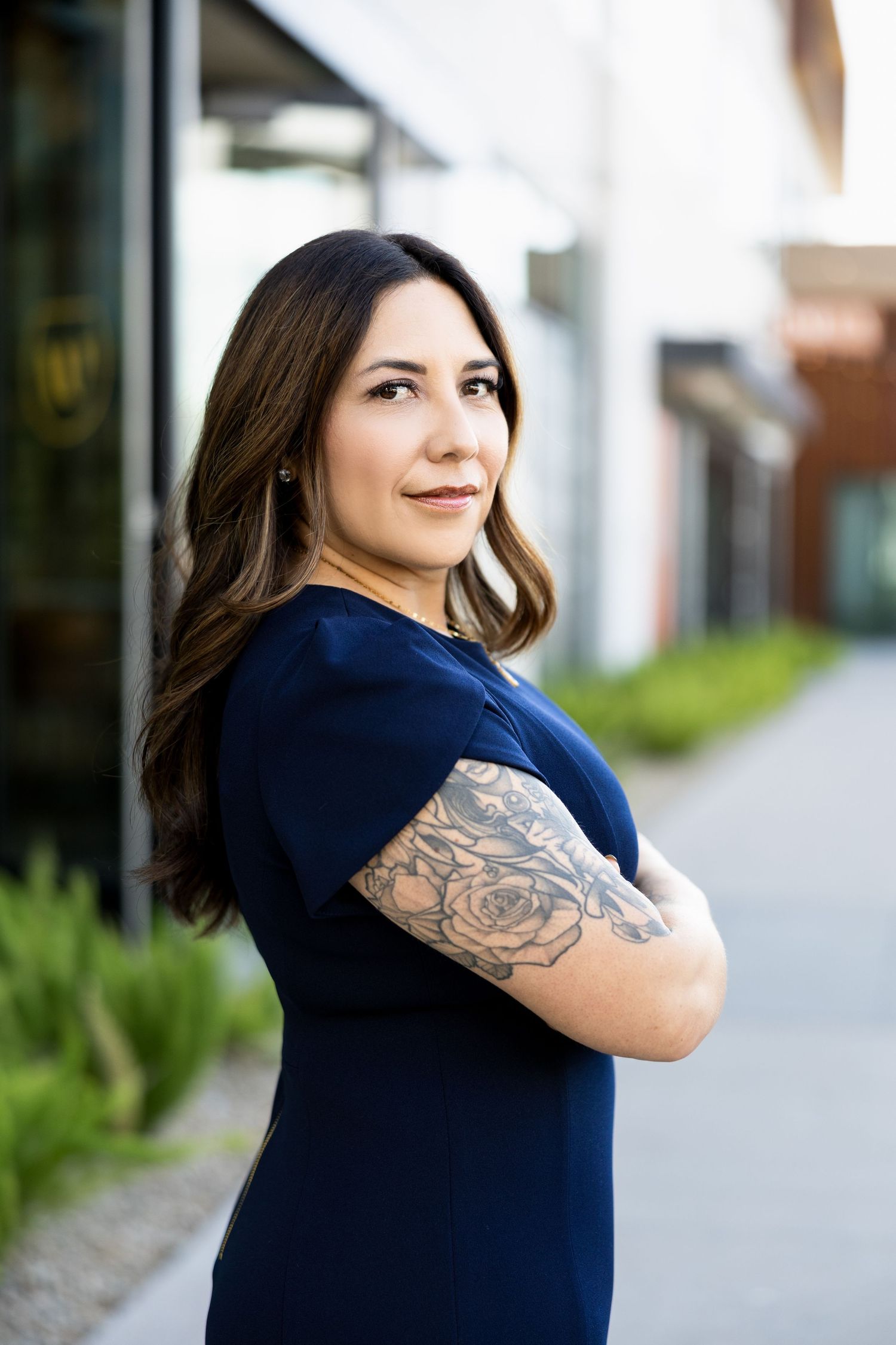 Woman in navy dress with floral arm tattoo stands confidently with arms crossed outdoors.