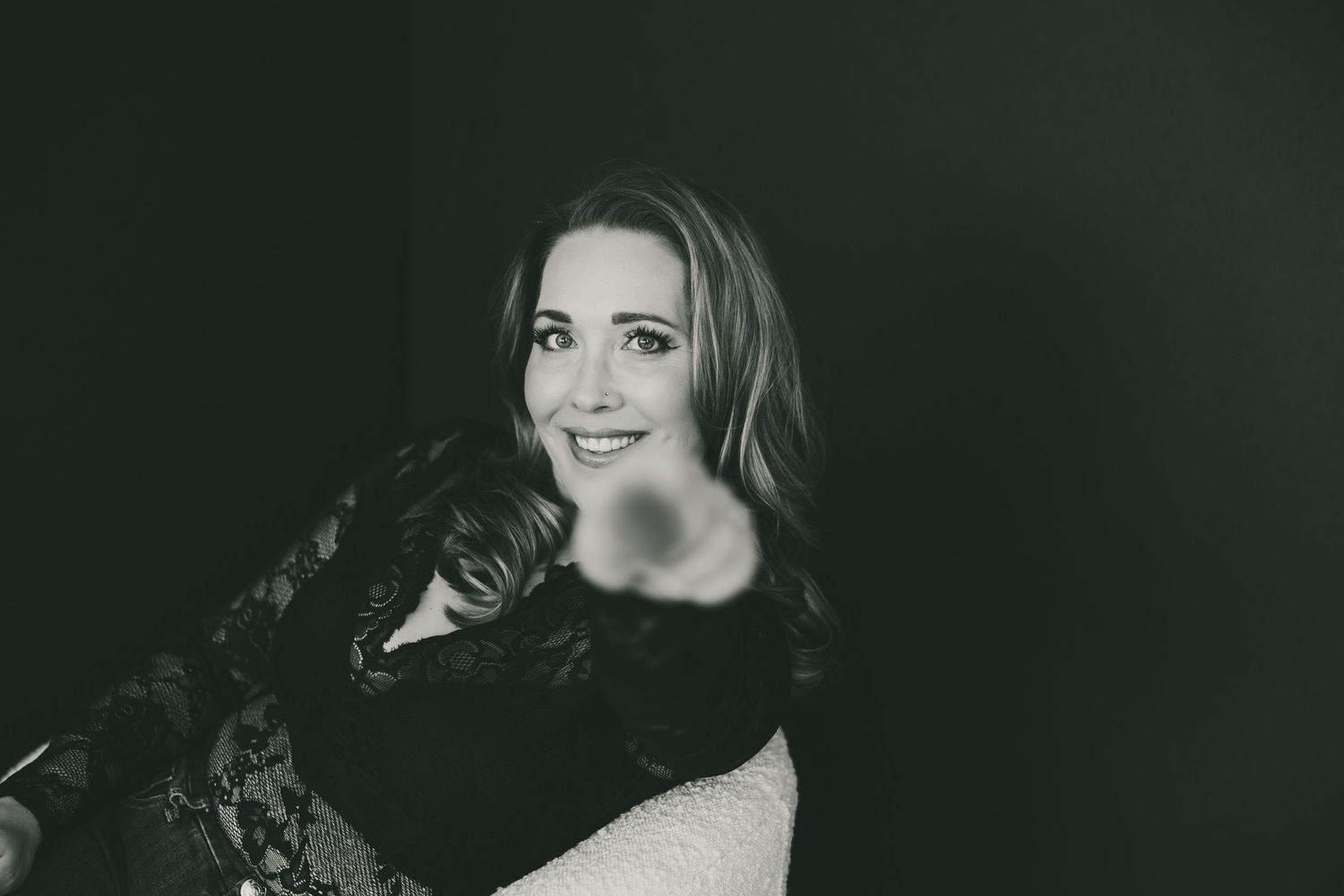 Smiling woman in black lace top gesturing toward camera in black and white portrait photo.
