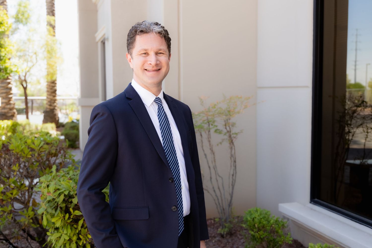 Professional man in navy suit and tie smiling outdoors near modern building with greenery.