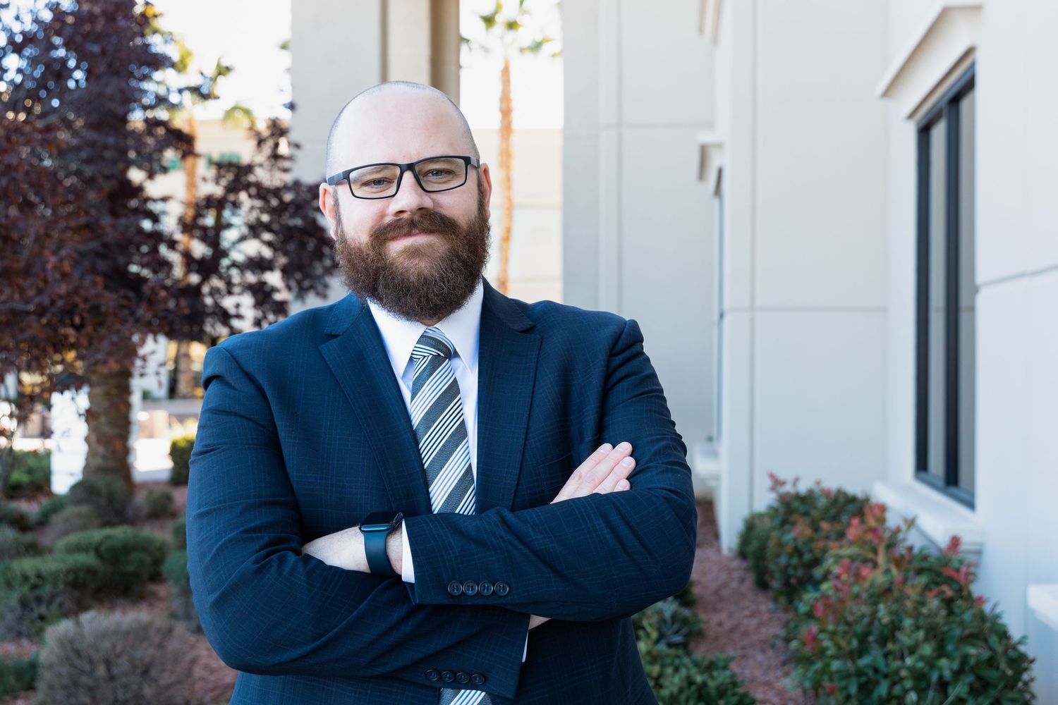 Professional businessman with beard and glasses poses confidently with arms crossed outdoors in navy suit.