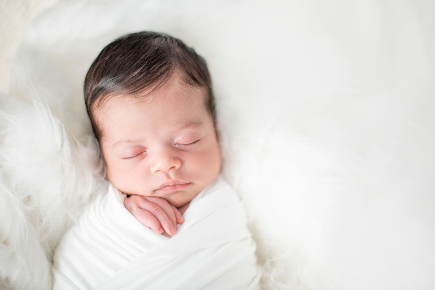 A sleeping newborn baby wrapped in soft white blankets rests peacefully against a white background.