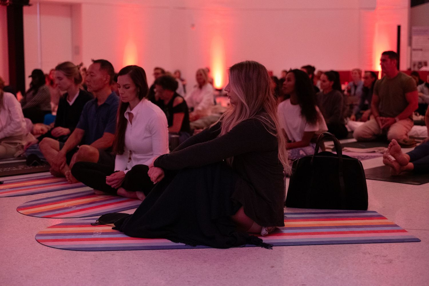 Group of people sitting cross-legged on colorful yoga mats during a meditation session in a pink-lit studio.