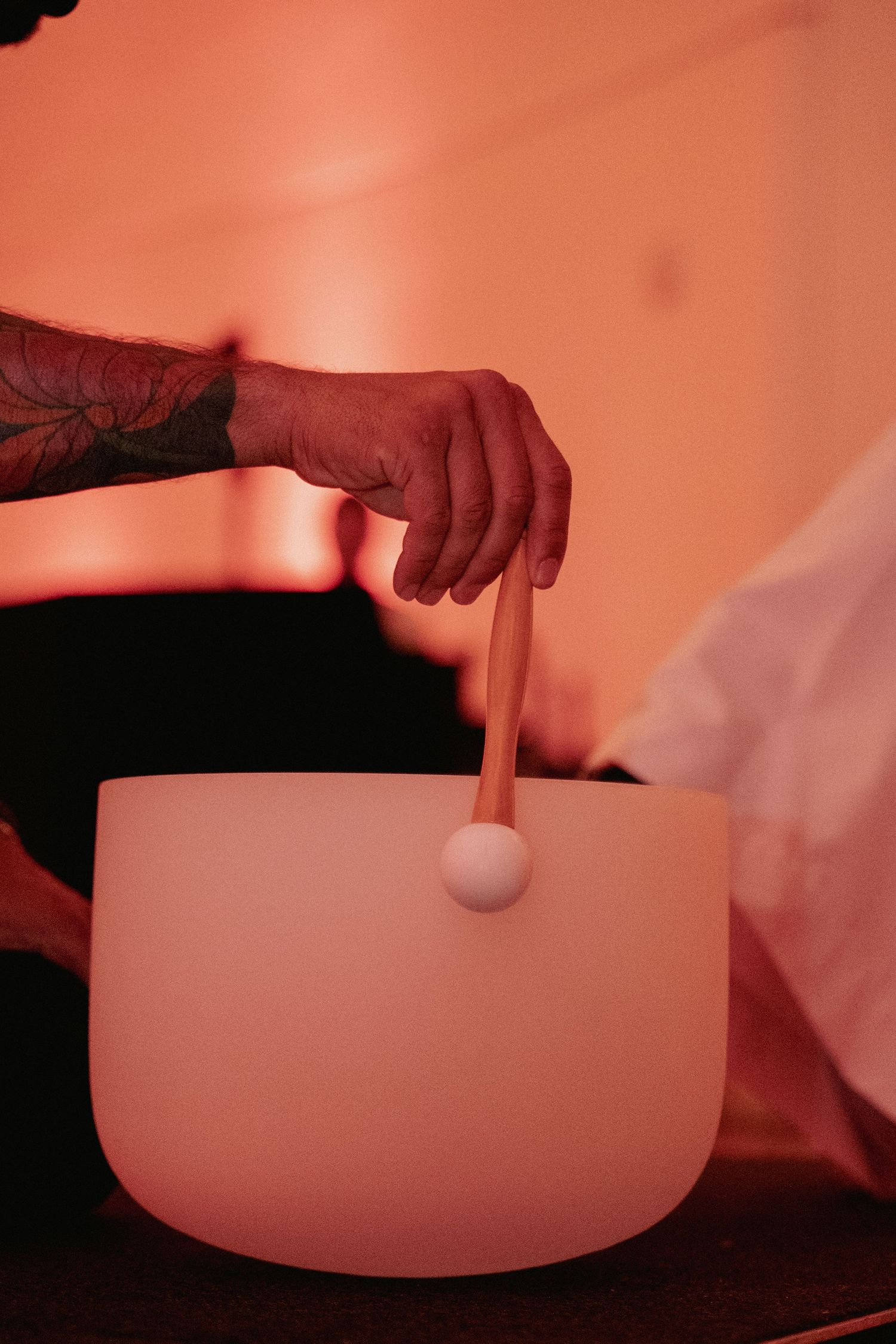 Tattooed hand playing a white crystal singing bowl with a mallet in warm orange ambient light.