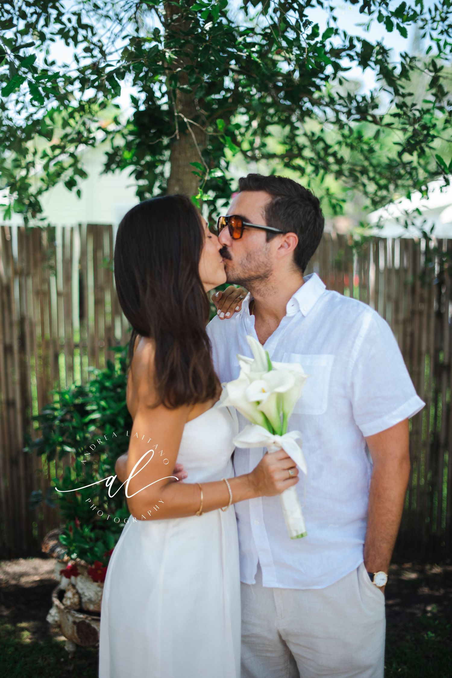 A romantic moment between a couple dressed in white against a bamboo fence backdrop.