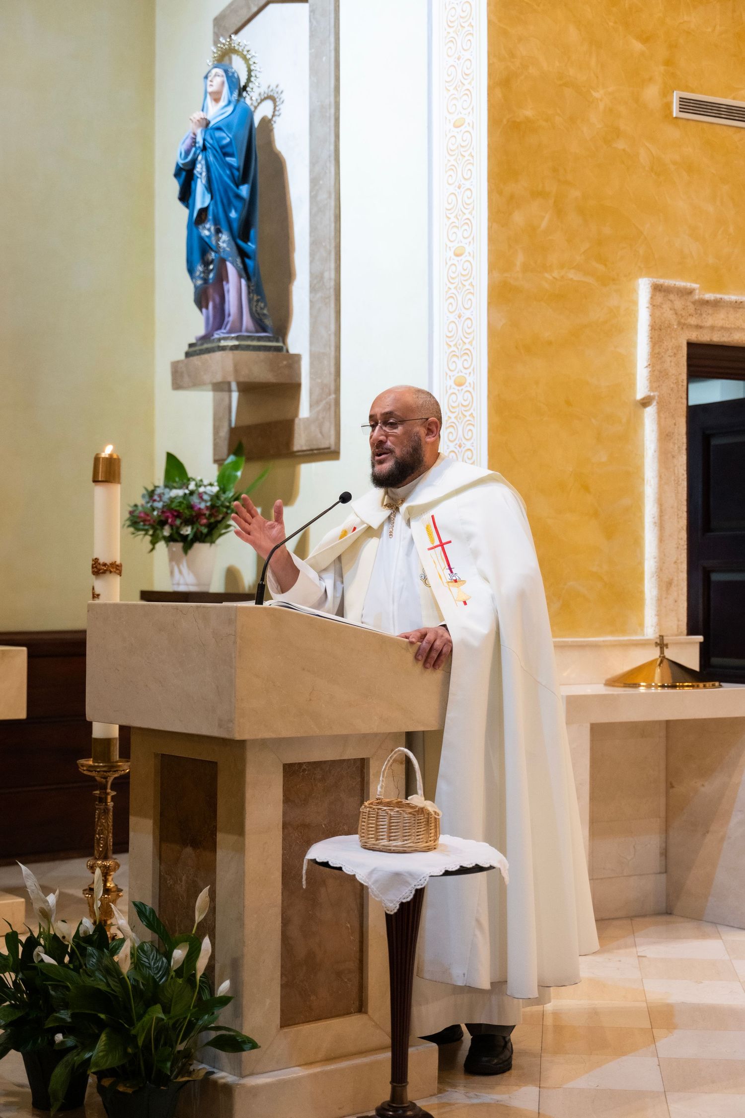 Catholic priest in white vestments delivers sermon at church pulpit with Virgin Mary statue in background.