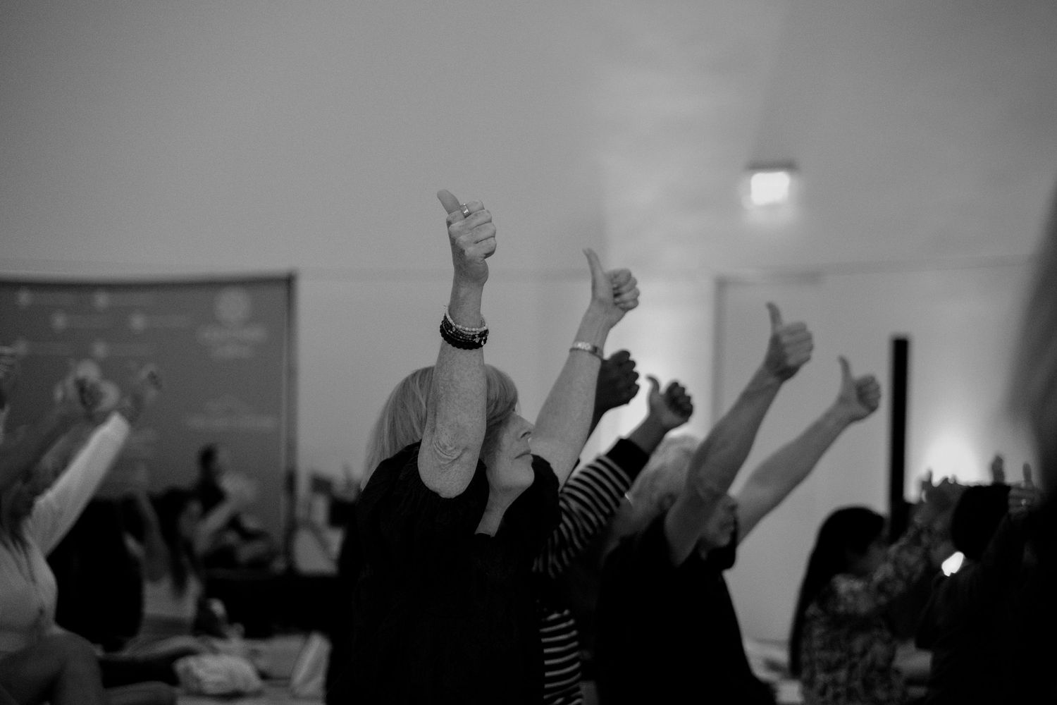 A group of people enthusiastically raise their thumbs up in a black and white photo at an indoor event.