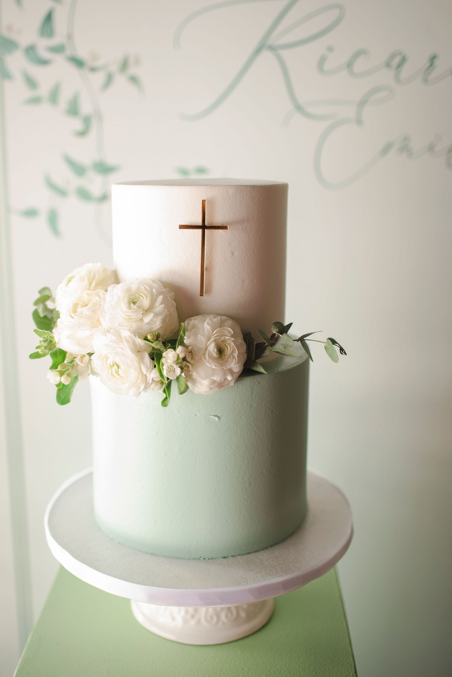 Elegant two-tier mint and white baptism cake decorated with fresh flowers and gold cross.