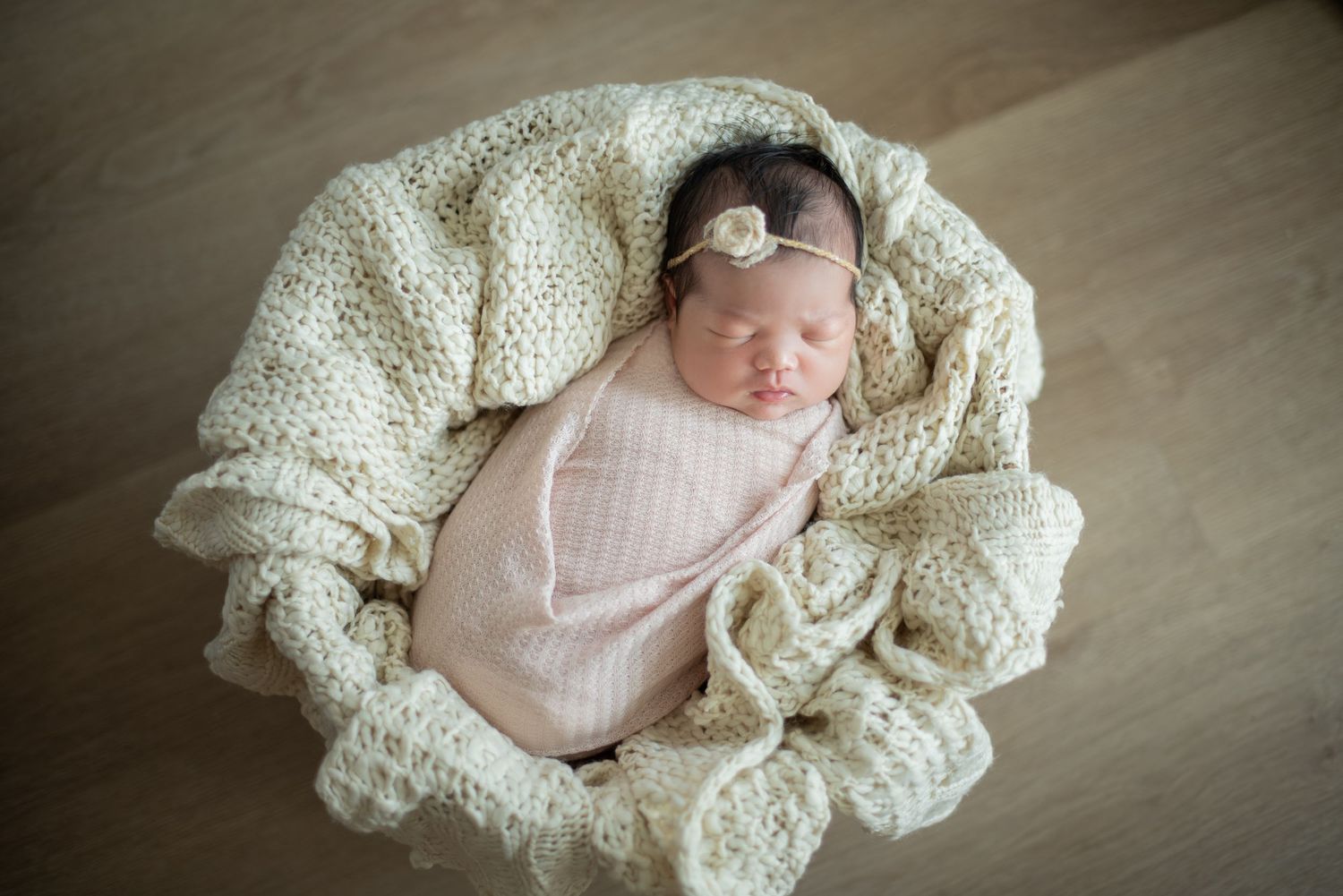 A sleeping newborn swaddled in soft pink fabric nestles in cream-colored knit blankets on a neutral backdrop.