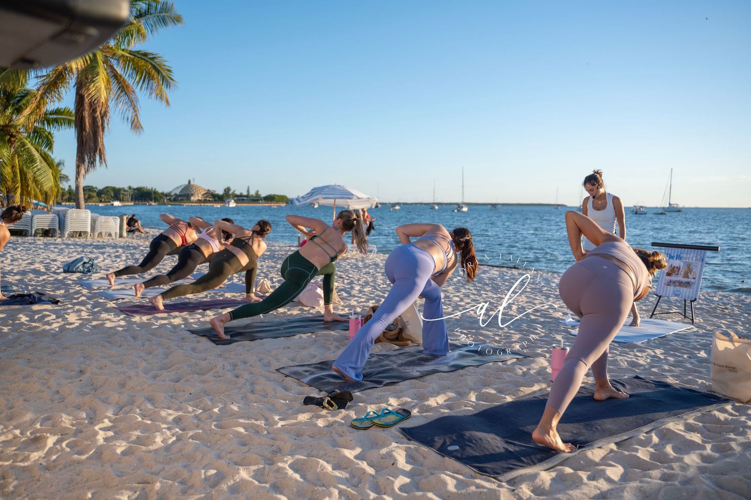 People practicing yoga poses together on a tropical beach at sunset with palm trees in the background.