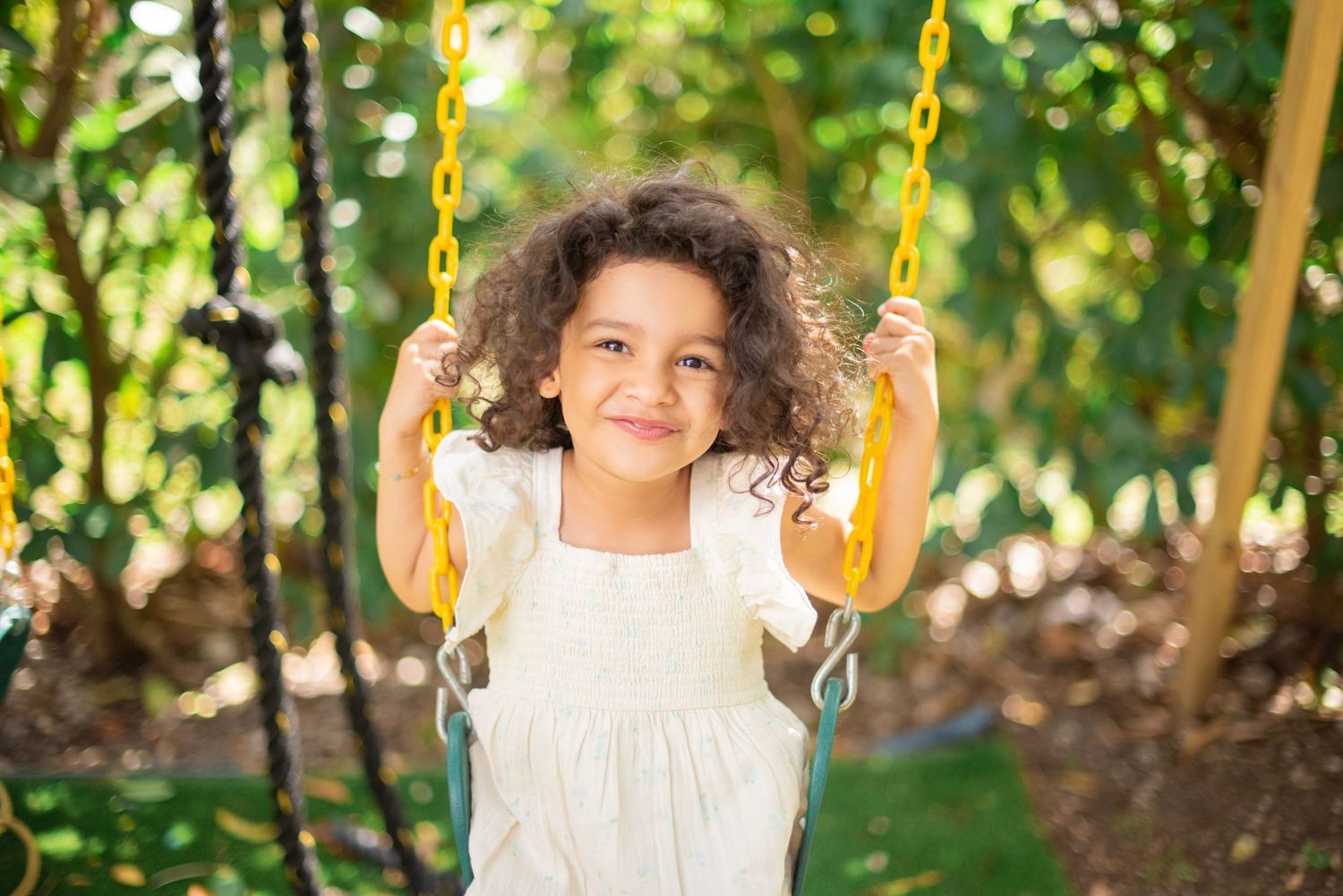A child in a white dress swings joyfully on yellow swings in a lush green garden setting.