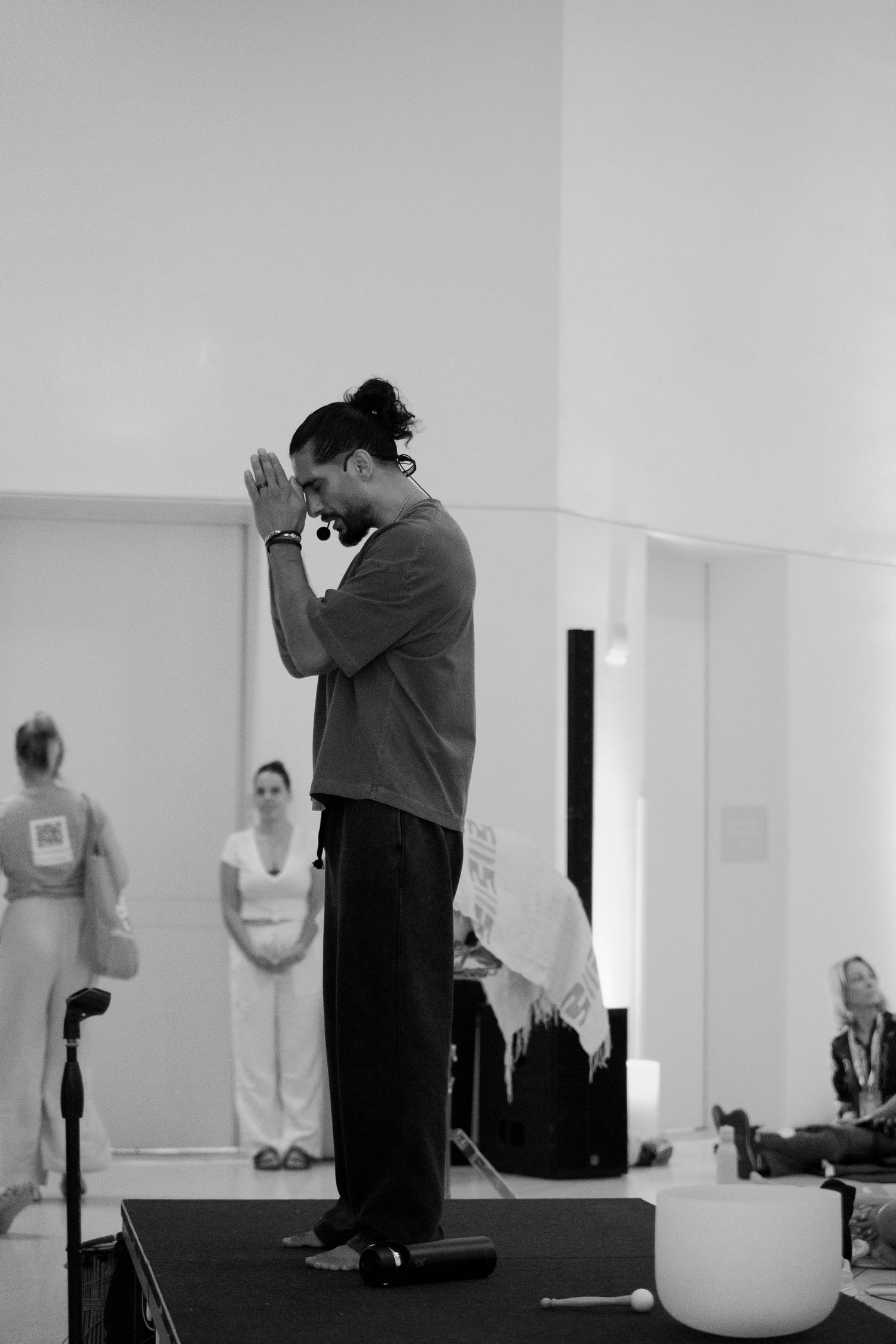 Man in prayer pose on stage at a yoga or wellness event with singing bowl nearby.