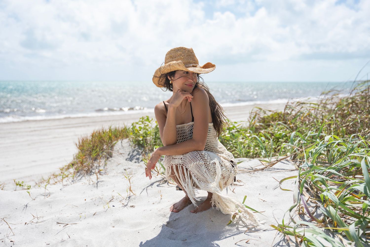 Person relaxing on a sandy beach with straw hat and ocean waves in the background.