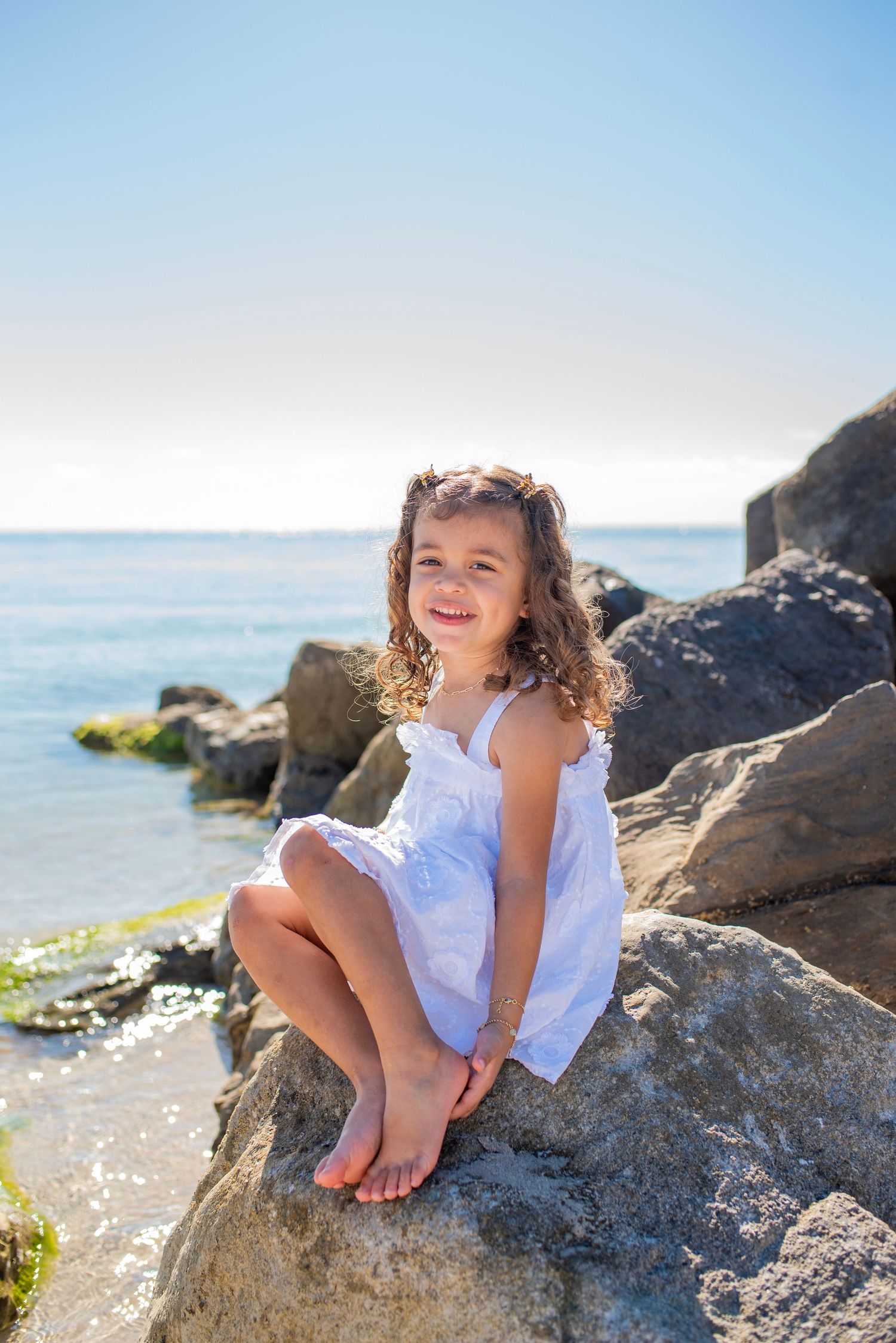 A child in a white dress sits playfully on coastal rocks by the ocean on a sunny summer day.