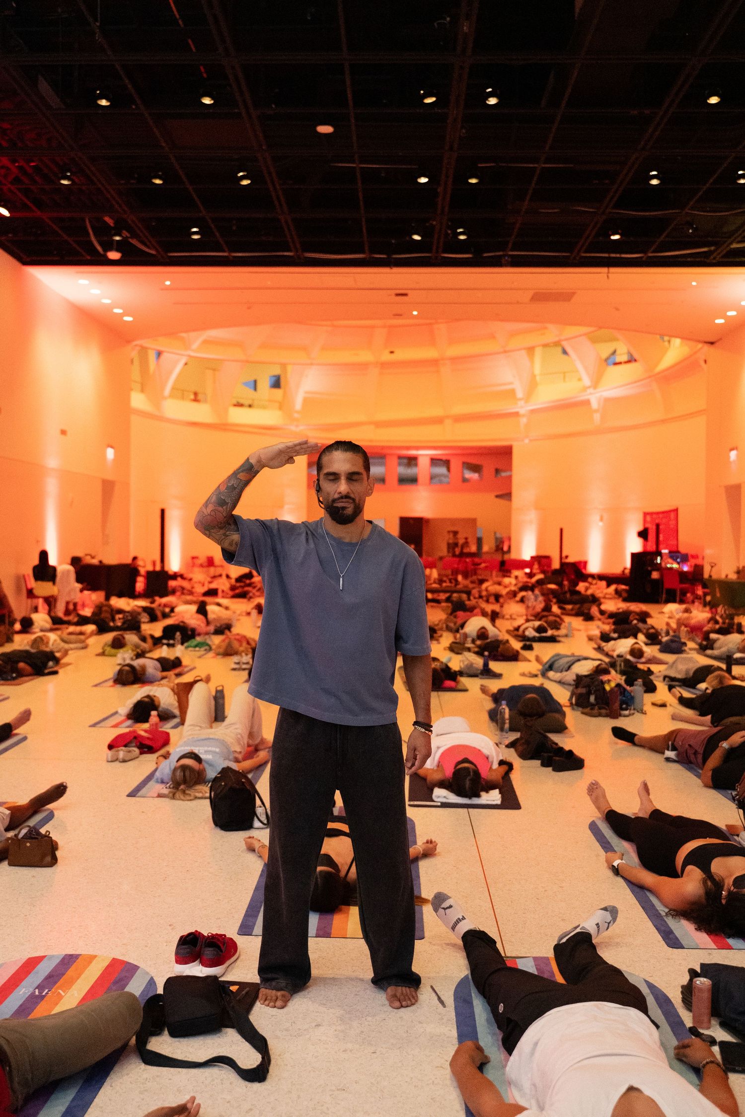 Tattooed yoga instructor stands among students lying on mats in a large warmly lit studio during a group class.