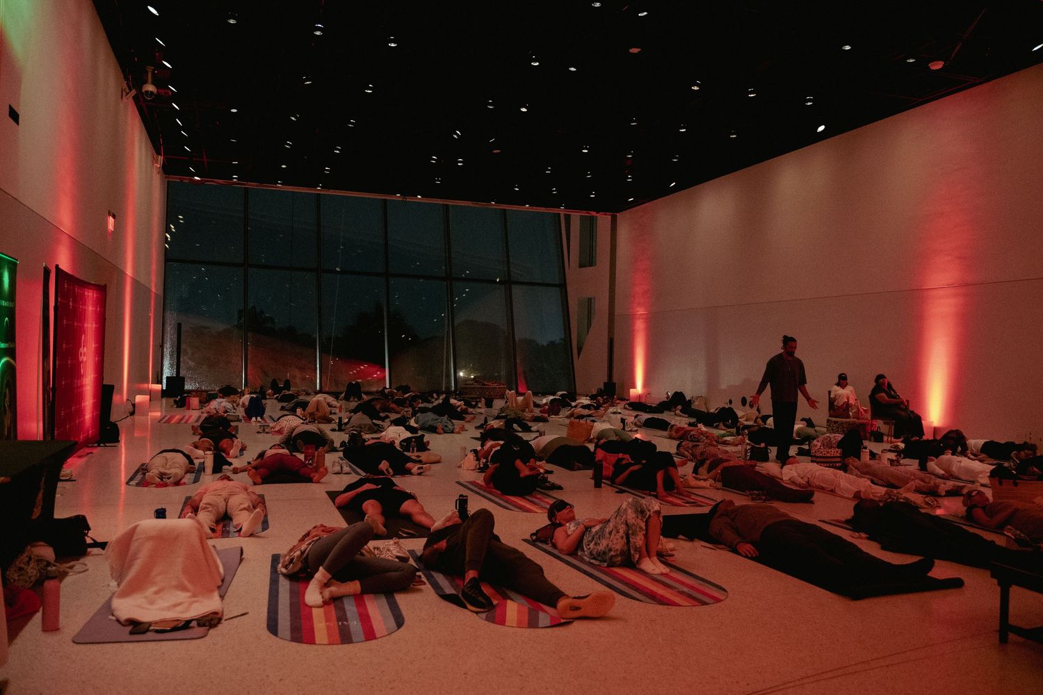 Group yoga or meditation session in a dimly lit modern hall with pink ambient lighting and floor mats.